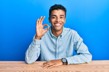 Genç, yakışıklı, Afro-Amerikalı, günlük giysiler içinde masada oturmuş gülümsüyor ve el ve parmaklarıyla 