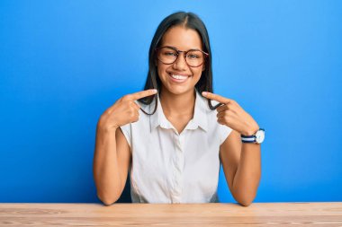 Güzel İspanyol kadın gündelik giysiler giyip masaya oturmuş gülümsüyor ve parmaklarıyla ve ağzıyla işaret ediyor. diş sağlığı kavramı. 