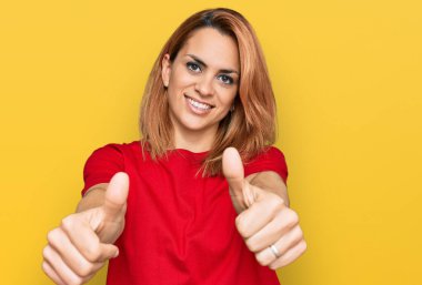 Latin kökenli genç bir kadın gündelik kırmızı t-shirt giyip olumlu bir jest yapmayı onaylıyor. Gülümsüyor ve başarı için seviniyor. kazanan jest. 