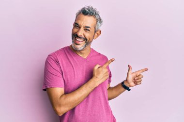 Orta yaşlı, gri saçlı, sıradan kıyafetler giyen, gülümseyen ve kameraya bakan, iki elini ve parmağını yana doğru gösteren bir adam.. 