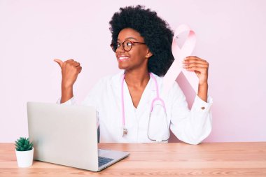 Genç Afro-Amerikan kadın doktor üniforması giyiyor elinde pembe kanser kurdelesi başparmağıyla yan tarafa bakıyor ve ağzı açık gülümsüyor. 