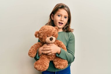 Küçük güzel kız oyuncak ayının yüzüne sarılmış, kuşkucu ve alaycı görünüyor, ağzı açık kalınca şaşırıyor. 