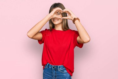 Gündelik kırmızı tişört giyen, elleri ve parmaklarıyla kalp şekli yapan, tabeladan bakıp gülümseyen beyaz bir kız. 