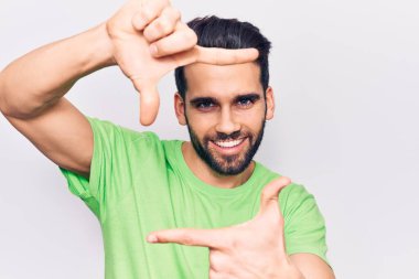 Sakallı, sıradan tişörtlü, gülümseyen, elleri ve parmakları gülen yüzlü genç yakışıklı bir adam. yaratıcılık ve fotoğrafçılık kavramı. 