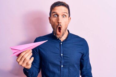 Pembe kağıt uçak tutan genç yakışıklı adam izole bir arka planda duruyor ve ağzı açık, korkmuş ve şaşkın bir şekilde şaşkınlık içinde, inançsız bir yüz.