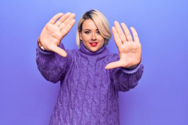 Güzel sarışın ve cüsseli bir kadın mor arka planda balıkçı yaka kazak giyiyor. El ve parmakları kullanarak çerçeve yapıyor. Kamera perspektifi kullanıyor.