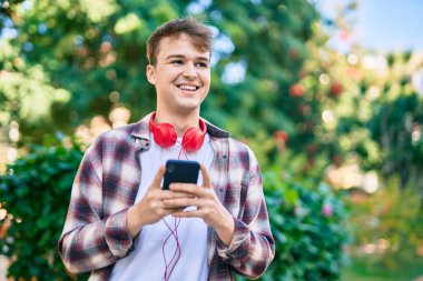 Parkta akıllı telefon ve kulaklık kullanarak gülümseyen beyaz bir genç..