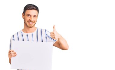 Elinde boş pankart tutan yakışıklı genç adam mutlu ve olumlu gülümsüyor, başparmağını kaldırıp mükemmel ve onay işareti yapıyor. 