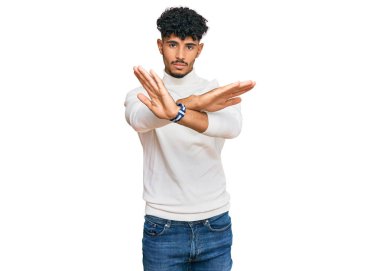 Kış süveteri giyen genç Arap adam kollarını kavuşturup, avuçlarını çaprazlayarak negatif işaret veriyor, kızgın surat yapıyor. 