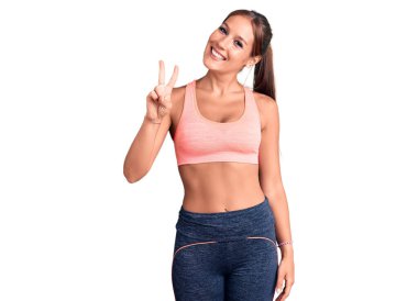 Genç ve güzel İspanyol kadın spor giysisi giyiyor. İki numarayı gösterip işaret ediyor. Kendinden emin ve mutlu bir şekilde gülümsüyor.. 