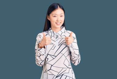 Gayri resmi tişört giyen genç, güzel Çinli bir kadın. Başarıyla olumlu jest yapıyor. Gülümsüyor ve mutlu. Neşeli ifade ve kazanan jest. 