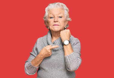 Kış süveteri giyen yaşlı, gri saçlı kadın saati, sabırsızlığı izlemek için acele ediyor ve rahat bir ifadeyle kameraya bakıyor. 