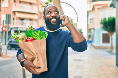Elinde süpermarketten alınmış kağıt torba sakallı Afrikalı Amerikalı adam telefonda konuşuyor.
