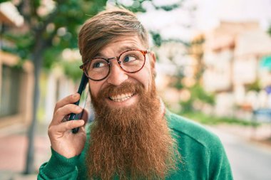 Kızıl saçlı genç İrlandalı adam mutlu bir şekilde gülümsüyor ve şehirdeki akıllı telefondan konuşuyor..