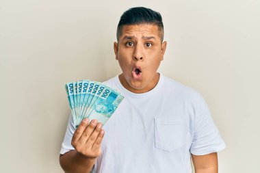 Genç Latin adam elinde 100 Brezilyalı gerçek banknot tutuyor. Şaşırmak için ağzı açık kalmış, korkmuş ve şaşırmış. İnanmayan yüz. 