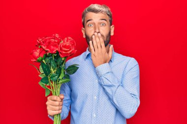 Yakışıklı, sarışın, romantik bir adam kırmızı arka planda gül buketi tutuyor. Ağzı elle kaplıyor, şok geçiriyor ve hata yapmaktan korkuyor. Şaşırtıcı ifade.