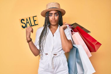 Saç örgülü, alışveriş torbası ve satış afişi taşıyan, kuşkucu ve sinirli genç Afro-Amerikalı kadın sorun yüzünden üzgün. negatif kişi. 