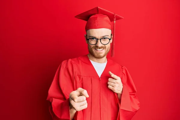 赤い卒業キャップと儀式用のローブを身に着けている若い赤毛の男は 幸せで面白い顔をしてカメラに指を指しています 良いエネルギーと振動は — ストック写真