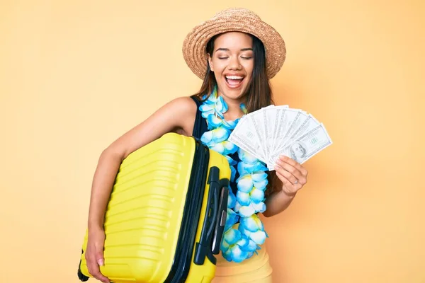 Young beautiful latin girl wearing hawaiian lei holding cabin bag and dollars banknotes smiling and laughing hard out loud because funny crazy joke.