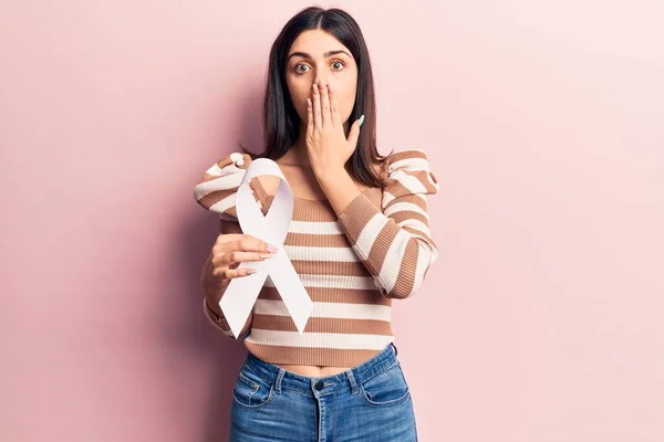 Young beautiful girl holding white cancer ribbon covering mouth with hand, shocked and afraid for mistake. surprised expression
