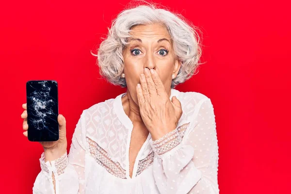 Senior grey-haired woman holding broken smartphone showing cracked screen covering mouth with hand, shocked and afraid for mistake. surprised expression