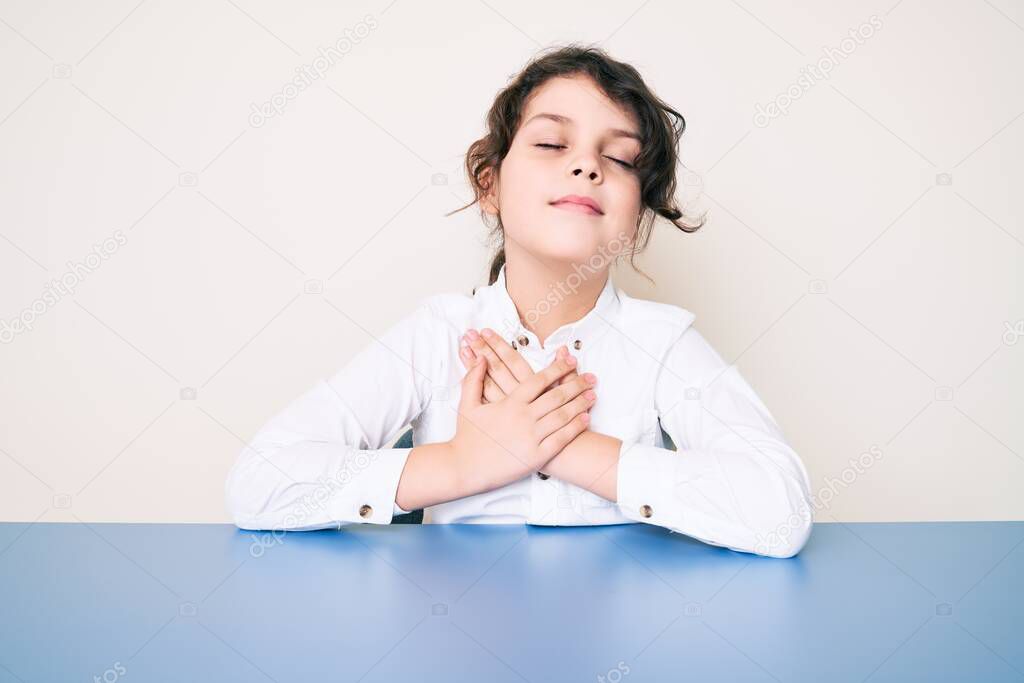 Cute hispanic child wearing casual clothes sitting on the table smiling with hands on chest, eyes closed with grateful gesture on face. health concept. 