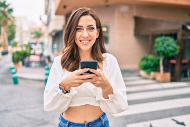 Genç İspanyol kadın şehirde akıllı telefon kullanarak mutlu bir şekilde gülümsüyor..