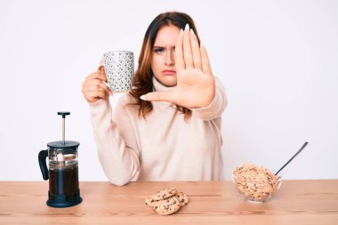 Genç, güzel, beyaz bir kadın masada oturmuş, eli açık kahvaltı yapıyor, dur işareti yapıyor, ciddi ve kendinden emin bir ifadeyle, savunma hareketi yapıyor. 