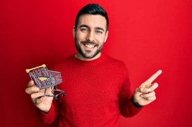 Genç İspanyol adam elinde küçük bir süpermarket arabası tutarken gülümsüyor mutlu bir şekilde elini ve parmağını kenarı gösteriyor. 