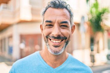 Orta yaşlı, kır saçlı, gülümseyen bir adam şehrin caddesinde mutlu bir şekilde yürüyor..