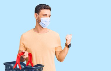 Alış veriş sepeti ve sağlık maskesi takmış yakışıklı genç adam baş parmağını yana kaldırıyor ve ağzı açık gülümsüyor. 