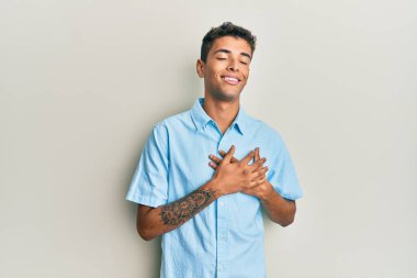 Genç, yakışıklı, Afro-Amerikalı sıradan kıyafetler giyen, elleri göğsünde, gözleri kapalı gülümseyen ve yüzünde minnet ifadesi olan bir adam. sağlık kavramı. 