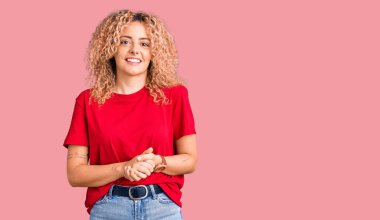 Kıvırcık saçlı, sıradan kırmızı tişörtlü, elleri çapraz parmaklı, gevşemiş ve neşeli gülümseyen genç sarışın kadın. Başarı ve iyimserlik 