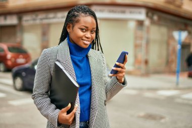 Genç Afrikalı Amerikalı iş kadını şehirde akıllı telefon kullanarak mutlu bir şekilde gülümsüyor..