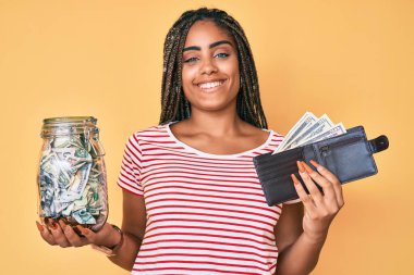 Tasarruf kavanozu ve cüzdanı olan mutlu ve havalı bir gülümsemeyle gülümseyen genç Afro-Amerikalı bir kadın. diş gösterme. 