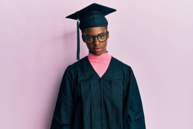 Mezuniyet şapkası ve tören cübbesi takan Afro-Amerikan genç kız yüzünde ciddi bir ifadeyle rahatlamış. Basit ve doğal bir şekilde kameraya bakıyorsun.. 