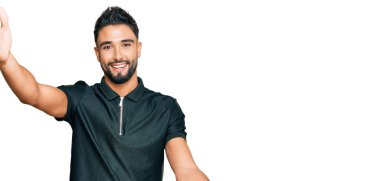 Sakallı genç bir adam spor kıyafetleri giymiş kameraya bakıyor ve sarılmak için kollarını açmış gülümsüyor. Mutluluğu kucaklayan neşeli bir ifade.. 