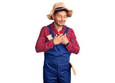 Yakışıklı Latin Amerikalı, giyimli, tamirci üniforması giyen, elleri göğsünde, gözleri kapalı gülümseyen ve yüzünde minnet ifadesi olan genç adam. sağlık kavramı. 