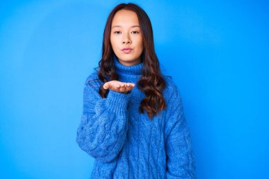 Young beautiful chinese girl wearing casual winter sweater looking at the camera blowing a kiss with hand on air being lovely and sexy. love expression. 