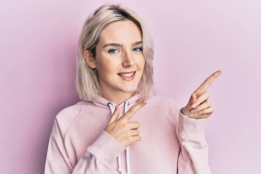 Sıradan kıyafetler giyen, gülümseyen ve kameraya bakan genç sarışın kız iki eliyle ve parmaklarıyla kenarı işaret ediyor.. 