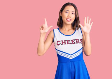 Genç, güzel Çinli kız ponpon kız üniforması giyiyor. Yedi numaralı parmaklarıyla işaret ediyor ve gülümsüyor. Kendinden emin ve mutlu.. 