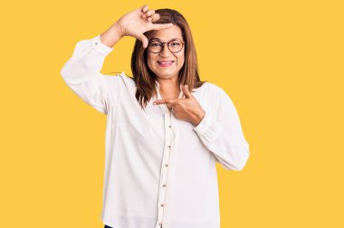 Orta yaşlı Latin kadın gündelik kıyafetler ve gözlük takıyor gülümsüyor elleri ve parmakları gülen yüzlü bir çerçeve yapıyor. yaratıcılık ve fotoğrafçılık kavramı. 