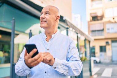 Orta yaşlı kel adam şehirde akıllı telefon kullanarak mutlu bir şekilde gülümsüyor..