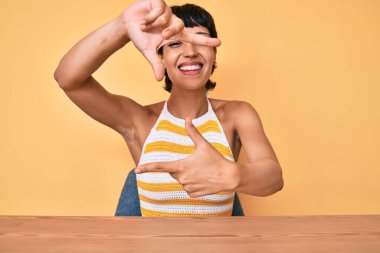 Masada oturan güzel esmer kadın sarı arka planda gülümsüyor ve mutlu yüzlü elleri ve parmakları olan bir çerçeve yapıyor. yaratıcılık ve fotoğrafçılık kavramı. 
