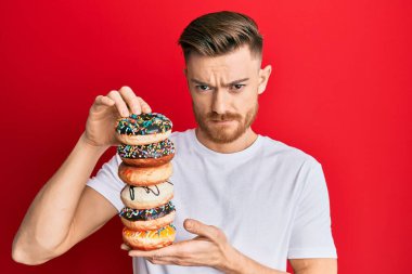 Genç kızıl saçlı adam elinde lezzetli renkli çörekler tutuyor şüpheci ve gergin, sorun yüzünden surat asıyor. negatif kişi. 