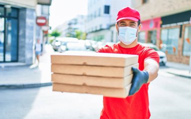 Üniforma giyen ve koronavirüs koruma maskesi takan genç teslimatçı. Kasaba caddesinde pizza kutularını teslim ediyorum..