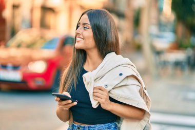 Genç İspanyol kız şehirde akıllı telefon kullanarak mutlu bir şekilde gülümsüyor..