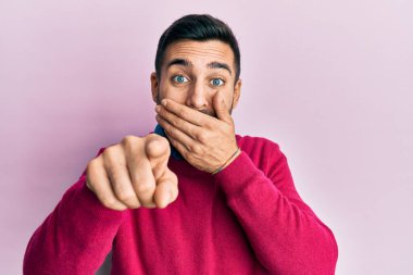 Gündelik kıyafetler giyen genç İspanyol adam sana gülüyor, eli ağzında kamerayı işaret ediyor, yüz ifadesi utanç verici. 