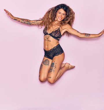 Genç, güzel, kıvırcık, dövmeli, iç çamaşırı giyen mutlu bir kadın. Yüzünde ve kollarında gülücüklerle pembe bir arka planda atlamak.