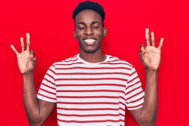 Genç Afro-Amerikan adam günlük kıyafetler giyip rahatlıyor ve gözleri kapalı gülümsüyor parmaklarıyla meditasyon hareketi yapıyor. yoga konsepti. 